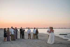 Wedding guests stand in a line along a sandy beach during golden hour with the ocean in the background.