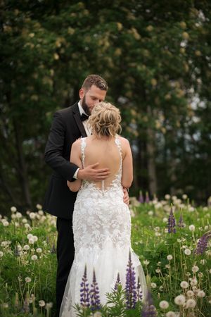 Bride and groom embrace in a wildflower meadow, her lace backless gown and his black tuxedo framed by lupines and trees.