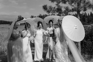 Black and white photo of a bride and bridesmaids holding parasols outdoors in a field with pine trees.