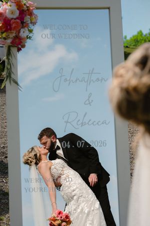 Bride and groom kissing in front of a wedding welcome mirror sign reading Johnathon and Rebecca 04.07.2026.