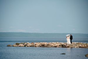 Bride and groom stand on rocky shoreline holding umbrella with calm blue ocean and distant hills backdrop.