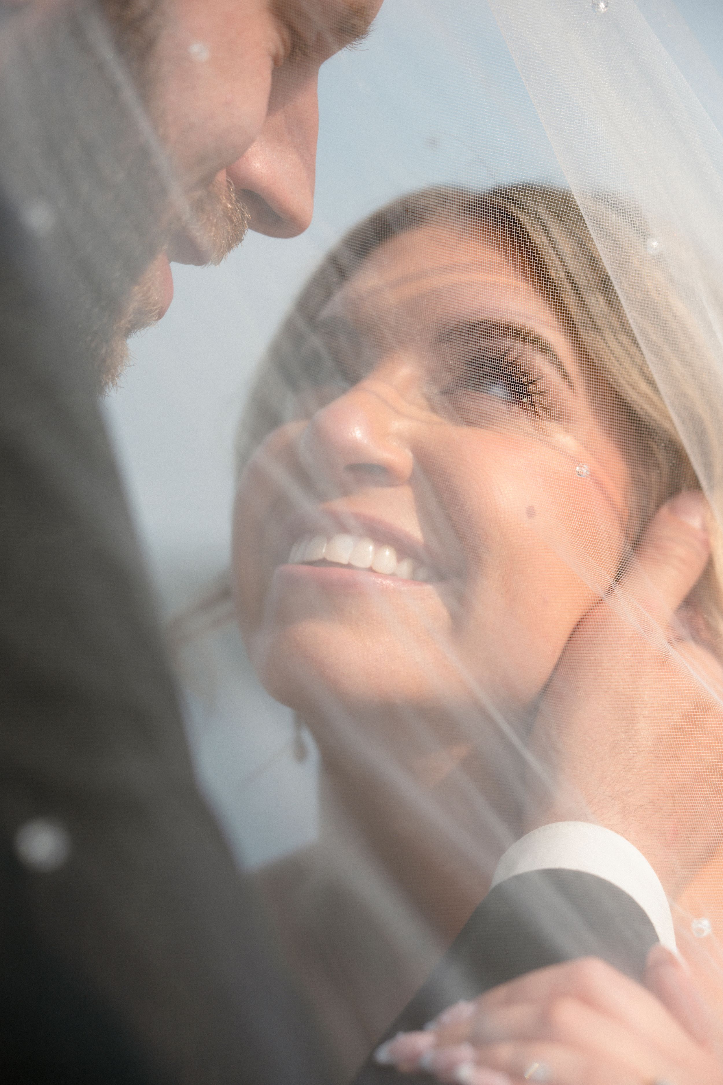 Bride smiling joyfully at groom under wedding veil, groom tenderly holding her face during romantic wedding moment.