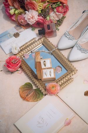 Elegant wedding flat lay with gold mirror tray, ring box, bridal heels, pink floral bouquet, and stationery on marble surface.