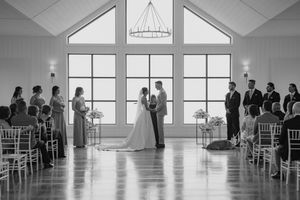 A black and white wedding ceremony in a bright chapel with white chairs and large windows.