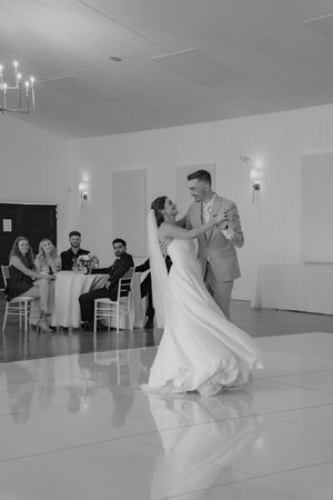 A couple shares their first dance at a wedding reception on a reflective white floor while guests watch from tables.