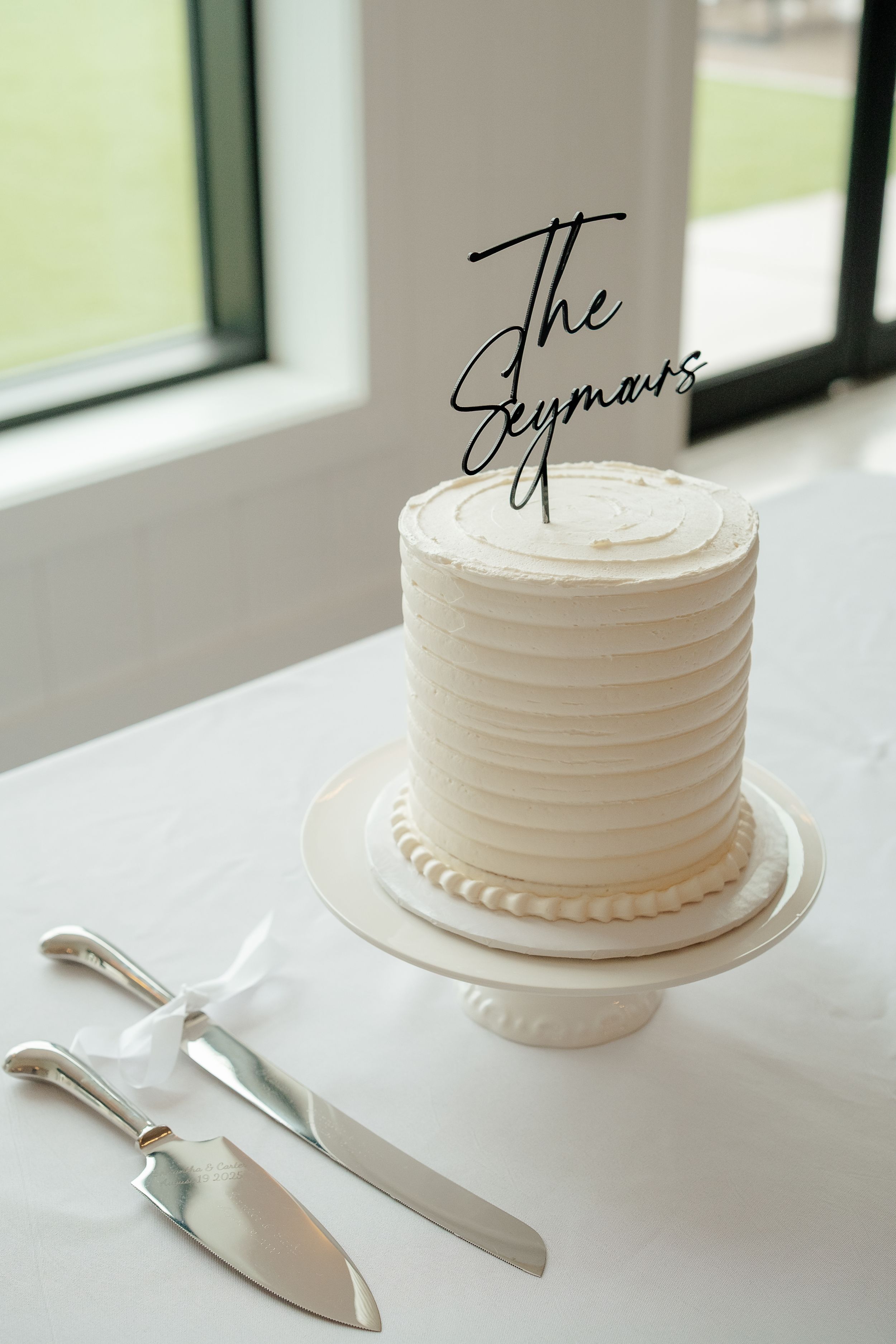 Elegant white wedding cake with textured buttercream frosting and black 'The Seymours' topper on a pedestal stand.