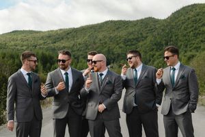 A group of groomsmen in gray suits and teal ties pose playfully in front of green rolling hills.