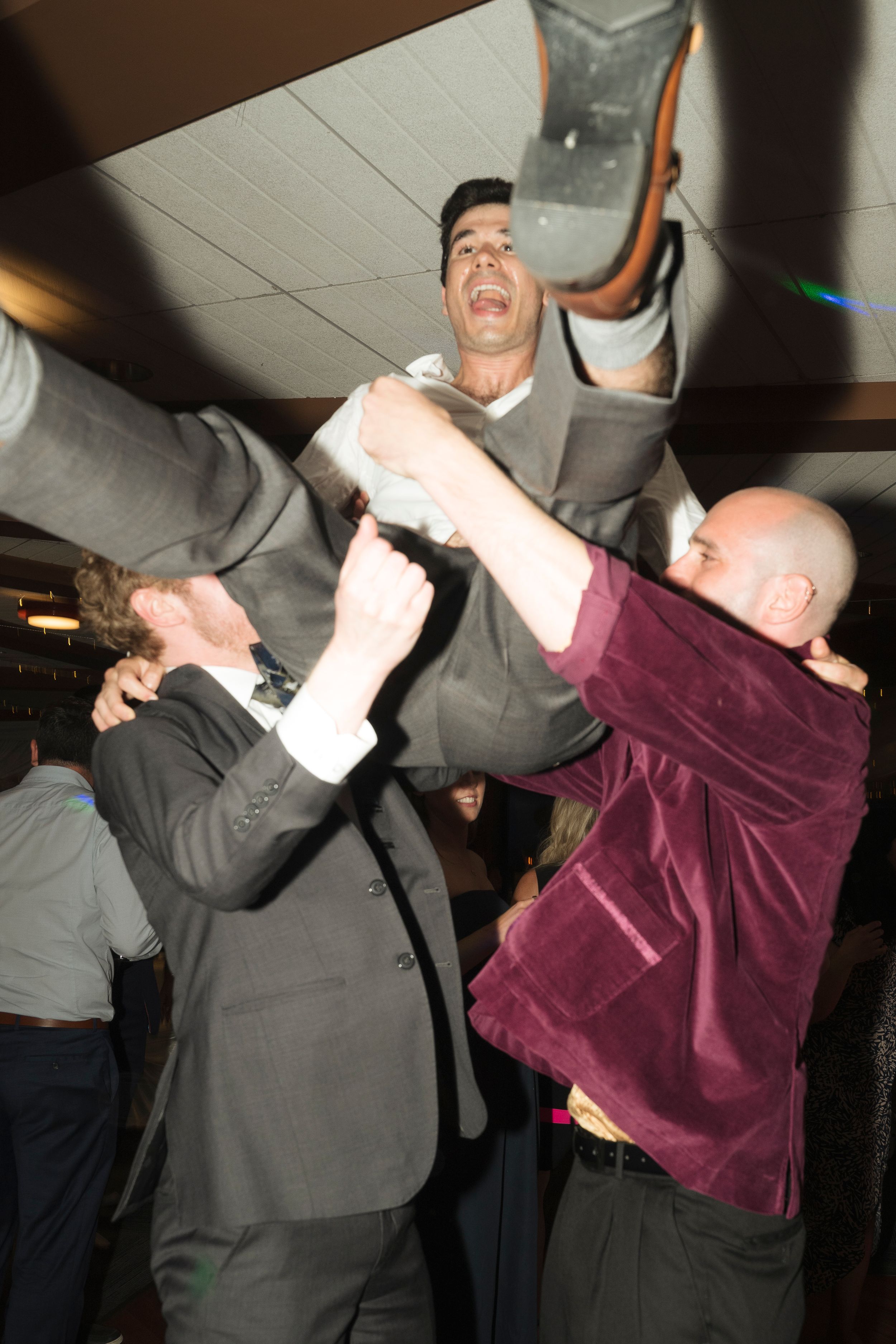 Men in suits lifting a celebrant at a lively party or wedding reception indoors.