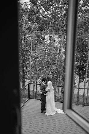 Black and white photo of bride and groom embracing on a wooden deck surrounded by lush trees.