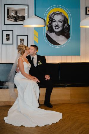 A bride in a flowing white gown kneels playfully on a wooden dance floor beneath retro-style wall decor.