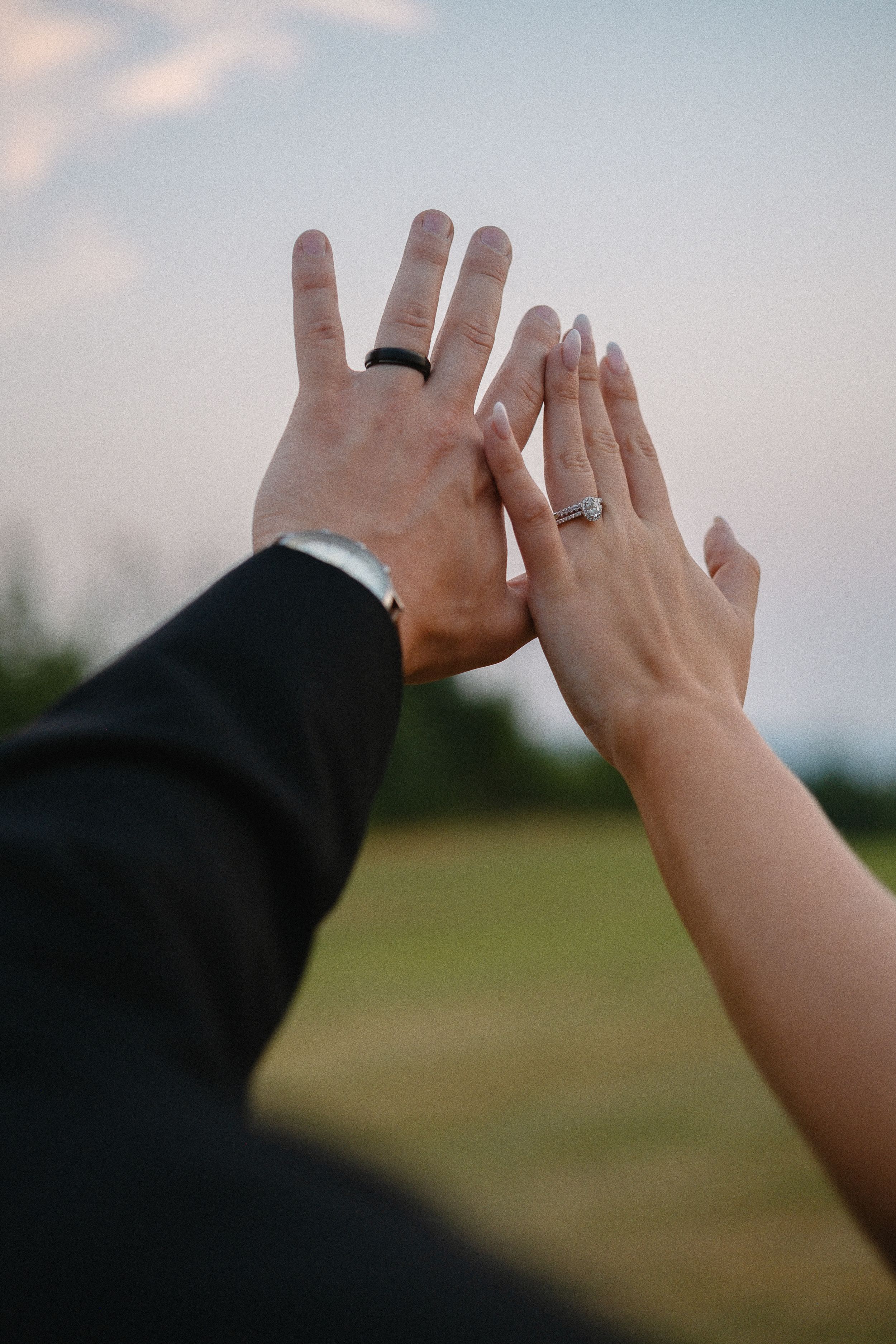 Two hands wearing wedding rings reach up to give each other a high five against a blurry outdoor background.