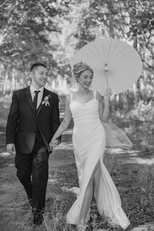 A couple shares a joyful moment during their wedding photoshoot in a wooded outdoor setting with a white parasol.