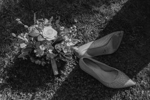 Black and white wedding details photo showing elegant bridal shoes and bouquet arranged on grass.