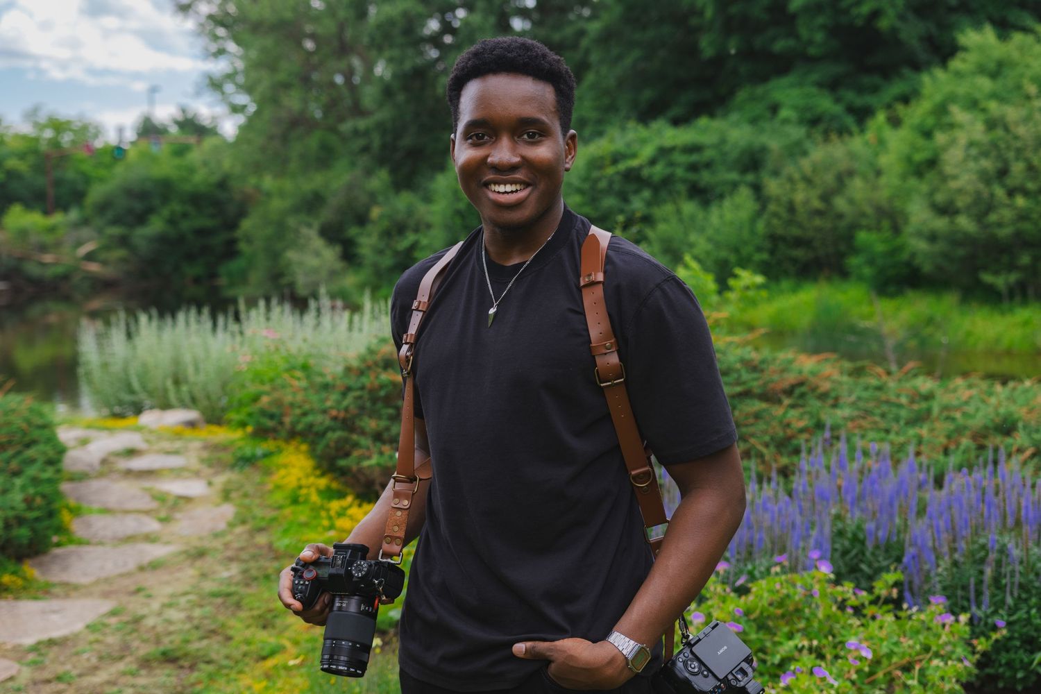 A photographer with camera gear stands on a garden path surrounded by purple flowers and greenery.