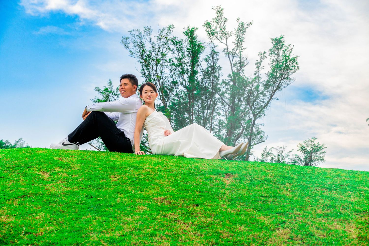Wedding couple relaxing on grassy hillside with trees in background.