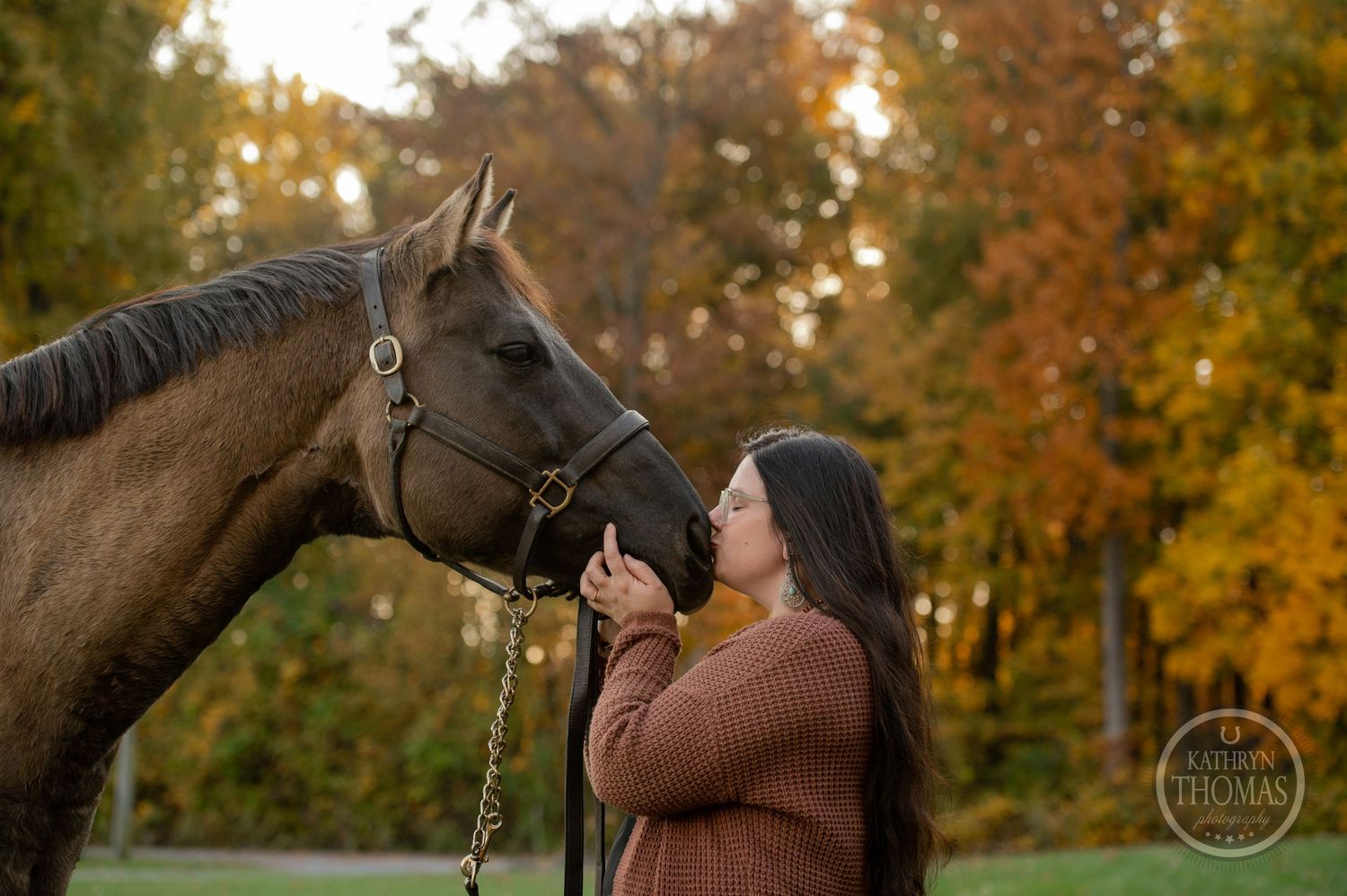 A horse and person share a tender moment against vibrant autumn foliage in a natural outdoor setting.