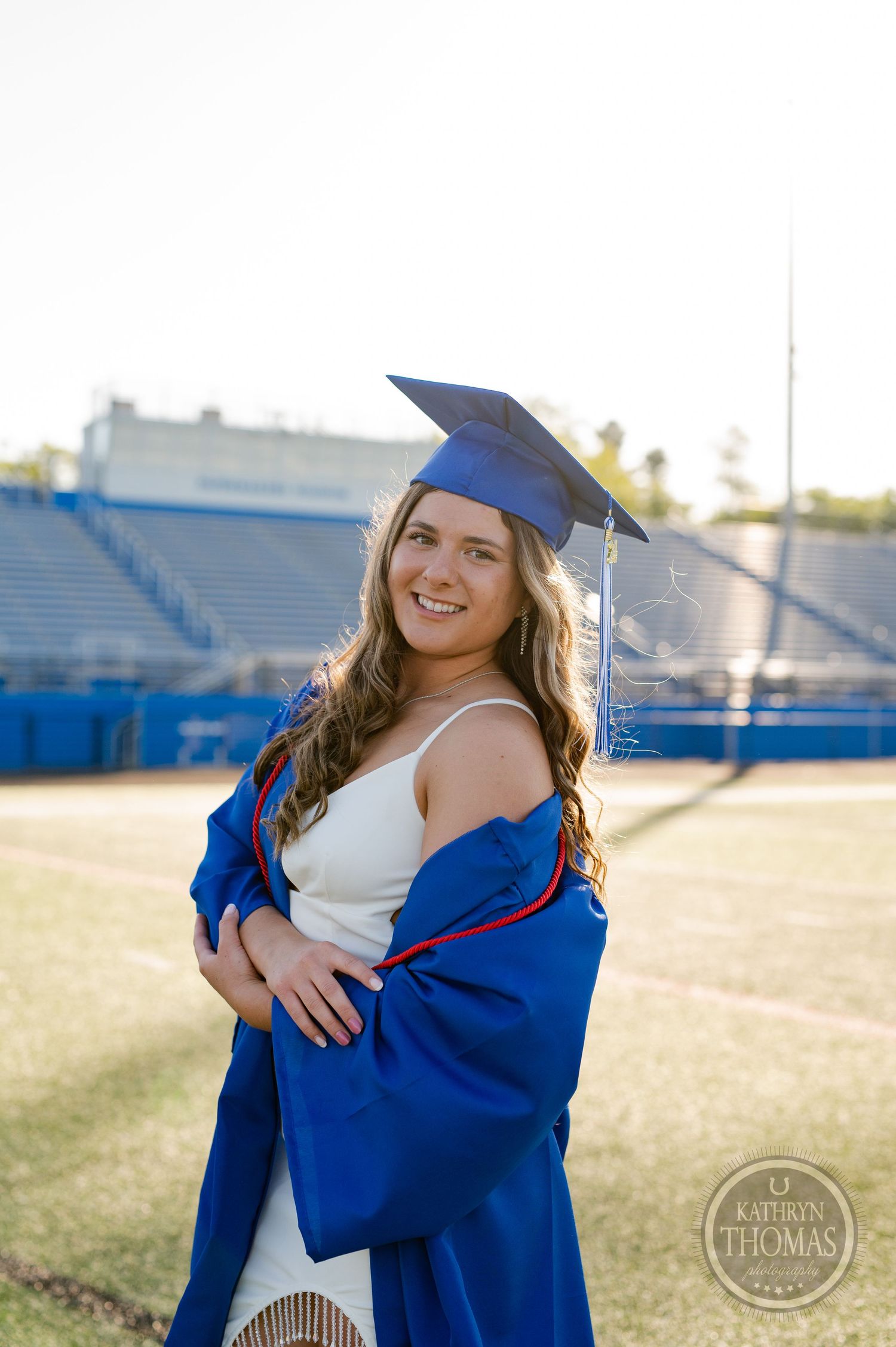 Graduate in white dress and blue cap and gown poses on sunny football field.