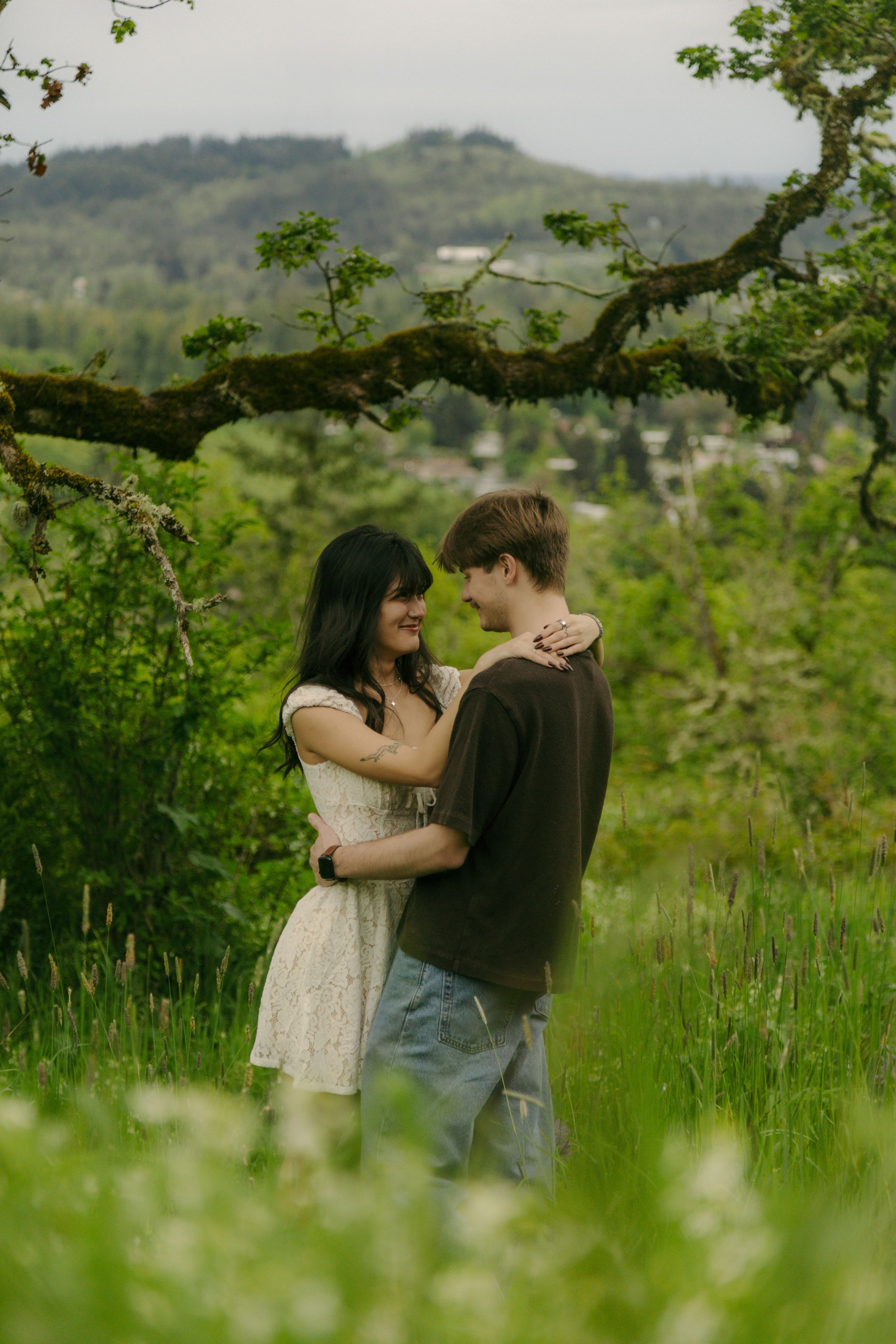 Couple embracing and smiling in a lush green field with a mossy tree branch and hills in the background.