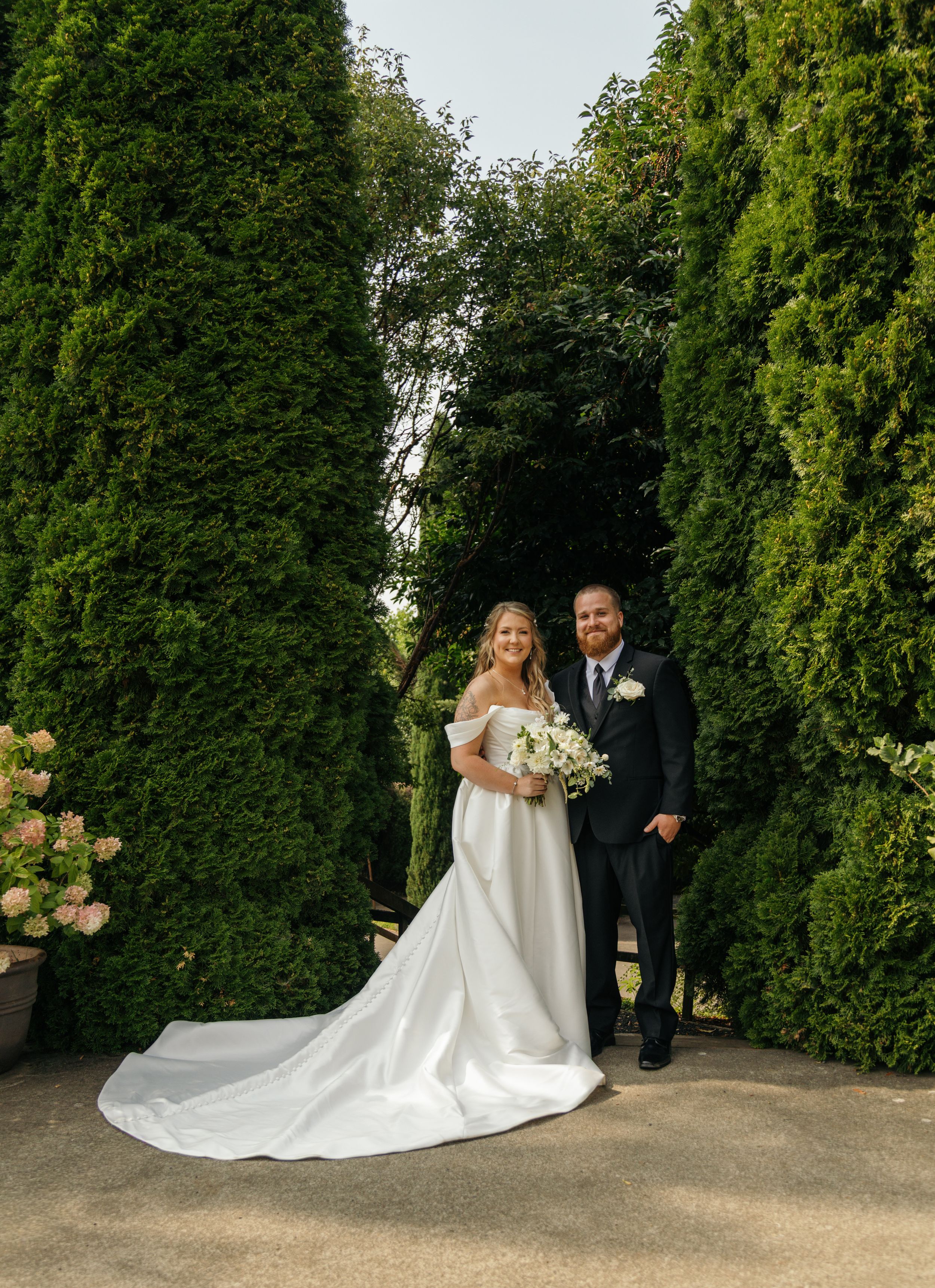 A wedding couple poses in a garden setting with lush green hedges and peach roses creating a romantic backdrop.