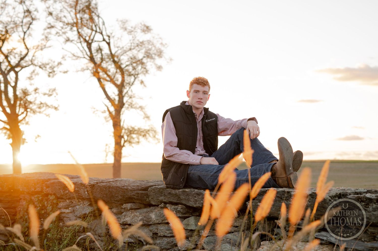 Person in casual attire sitting on stone wall during sunset with wheat grass in foreground.