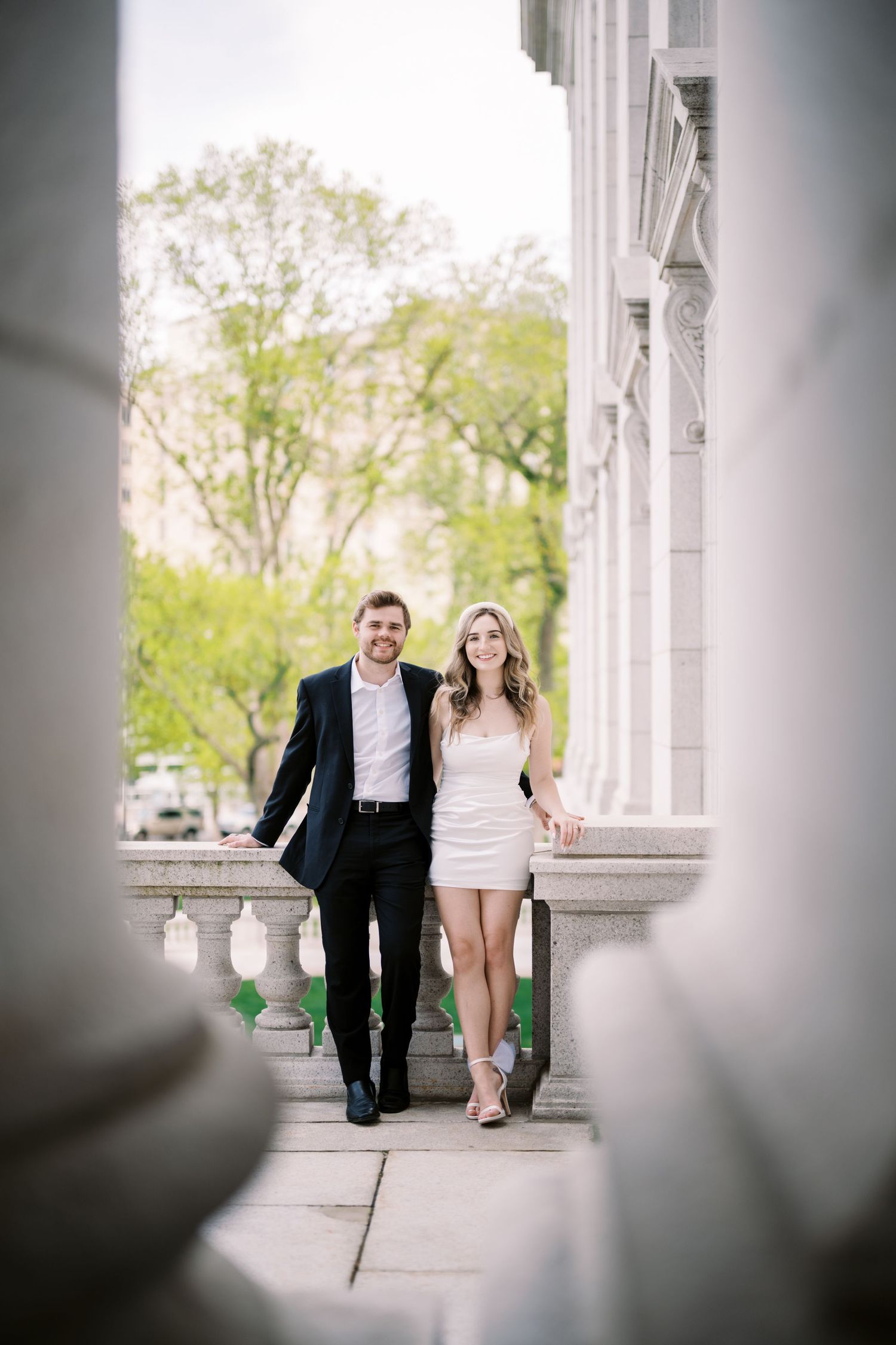 A couple in formal attire stands on white marble steps framed between stone columns on a spring day.
