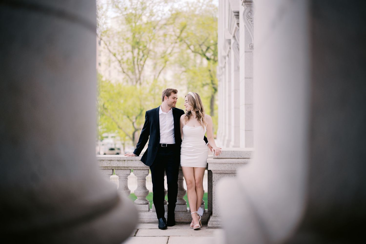 A romantic couple in formal attire walk together through elegant stone columns on a bright spring day.