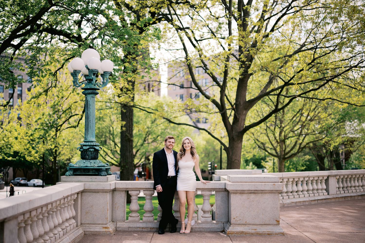 A couple poses on an ornate stone bridge with decorative lamp posts in a green park filled with trees.