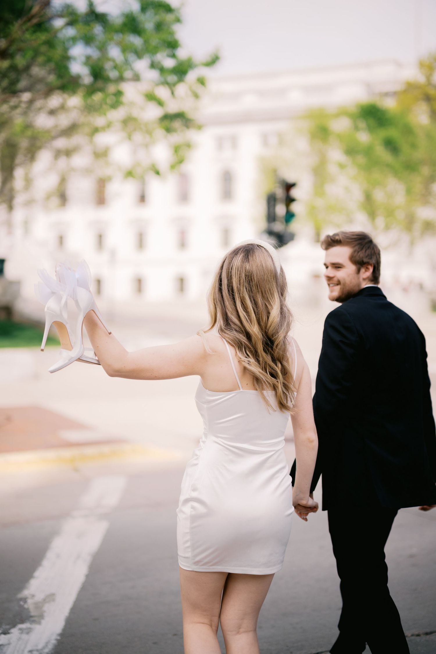 A couple in formal attire walks down a city street, with the person in white playfully raising their hand while walking.
