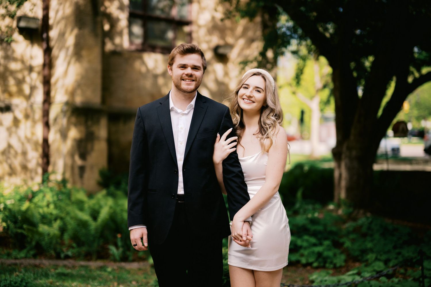 A couple in formal attire smiles while standing together in a lush garden setting on a sunny day.