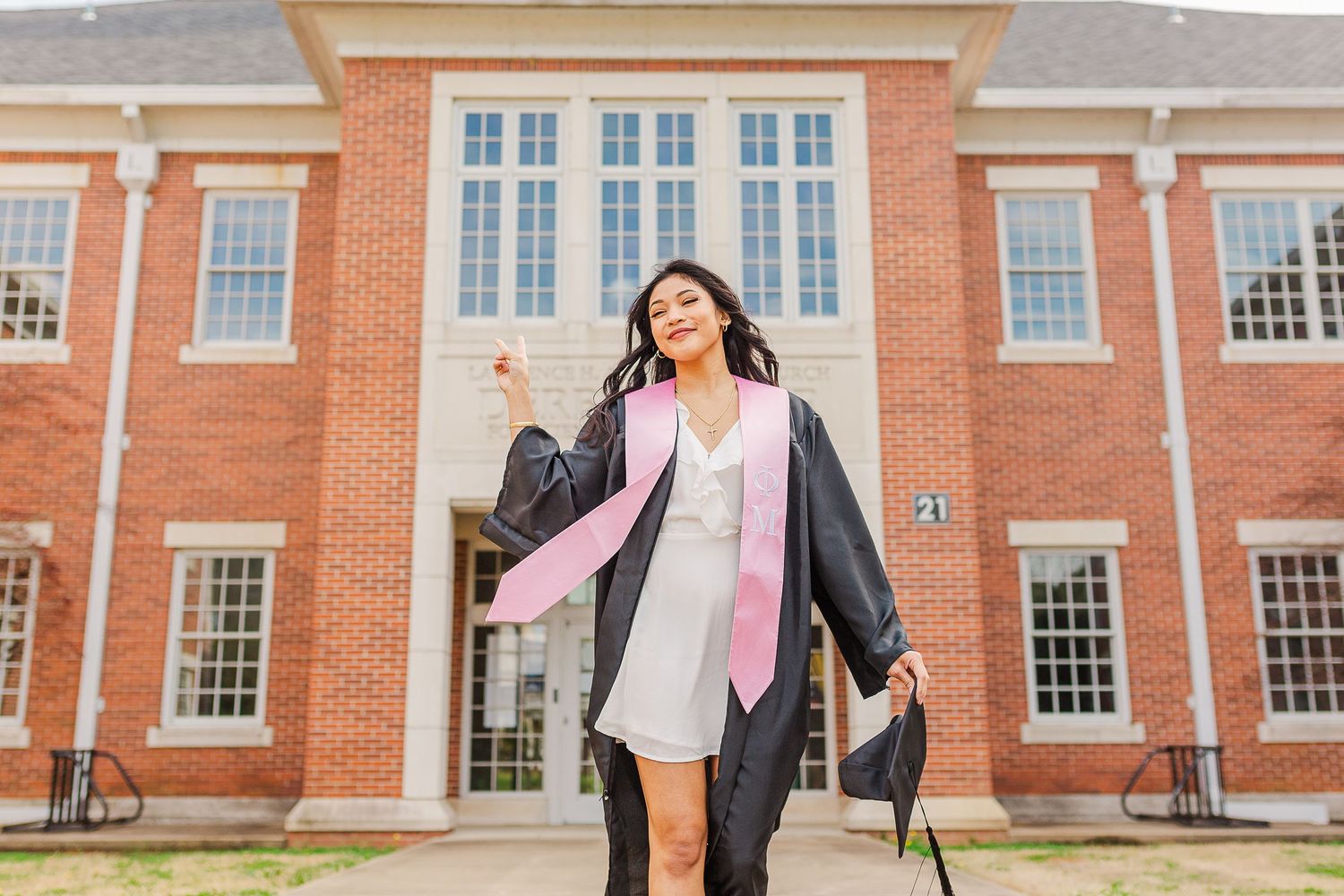 Graduate celebrates in cap and gown with pink graduation stole in front of red brick university building.