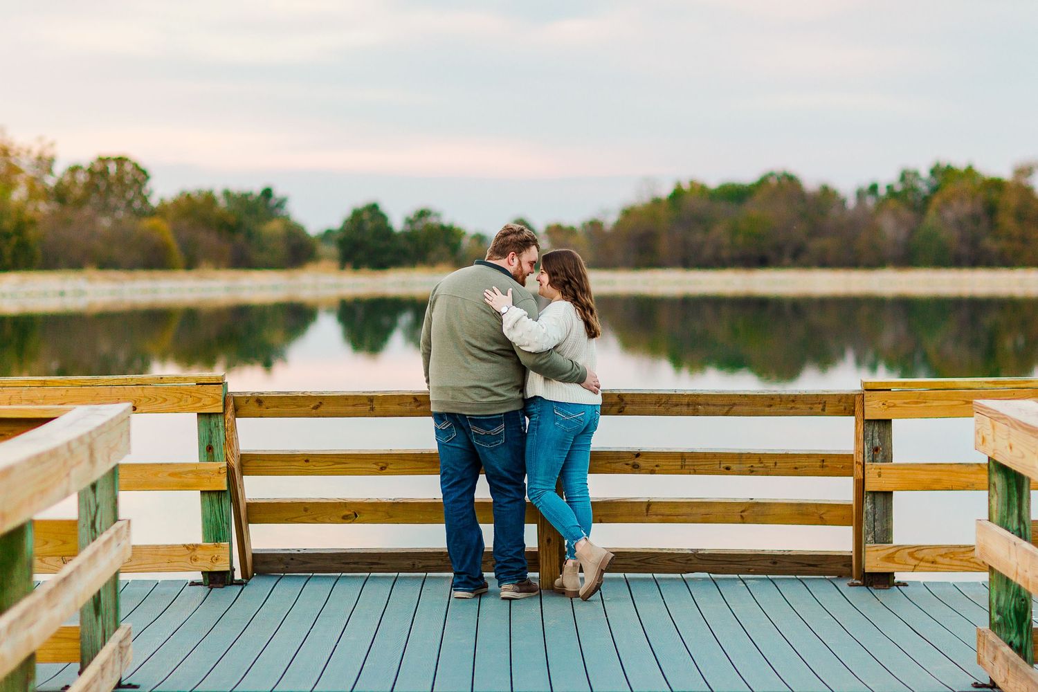 Couple embraces on a wooden dock overlooking a calm lake at sunset with autumn trees in background.