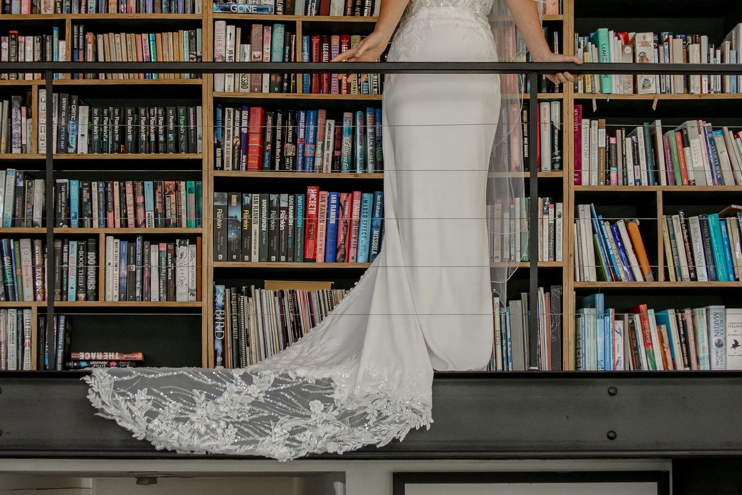 Long white wedding dress with lace train displayed against floor-to-ceiling library bookshelves in elegant setting.
