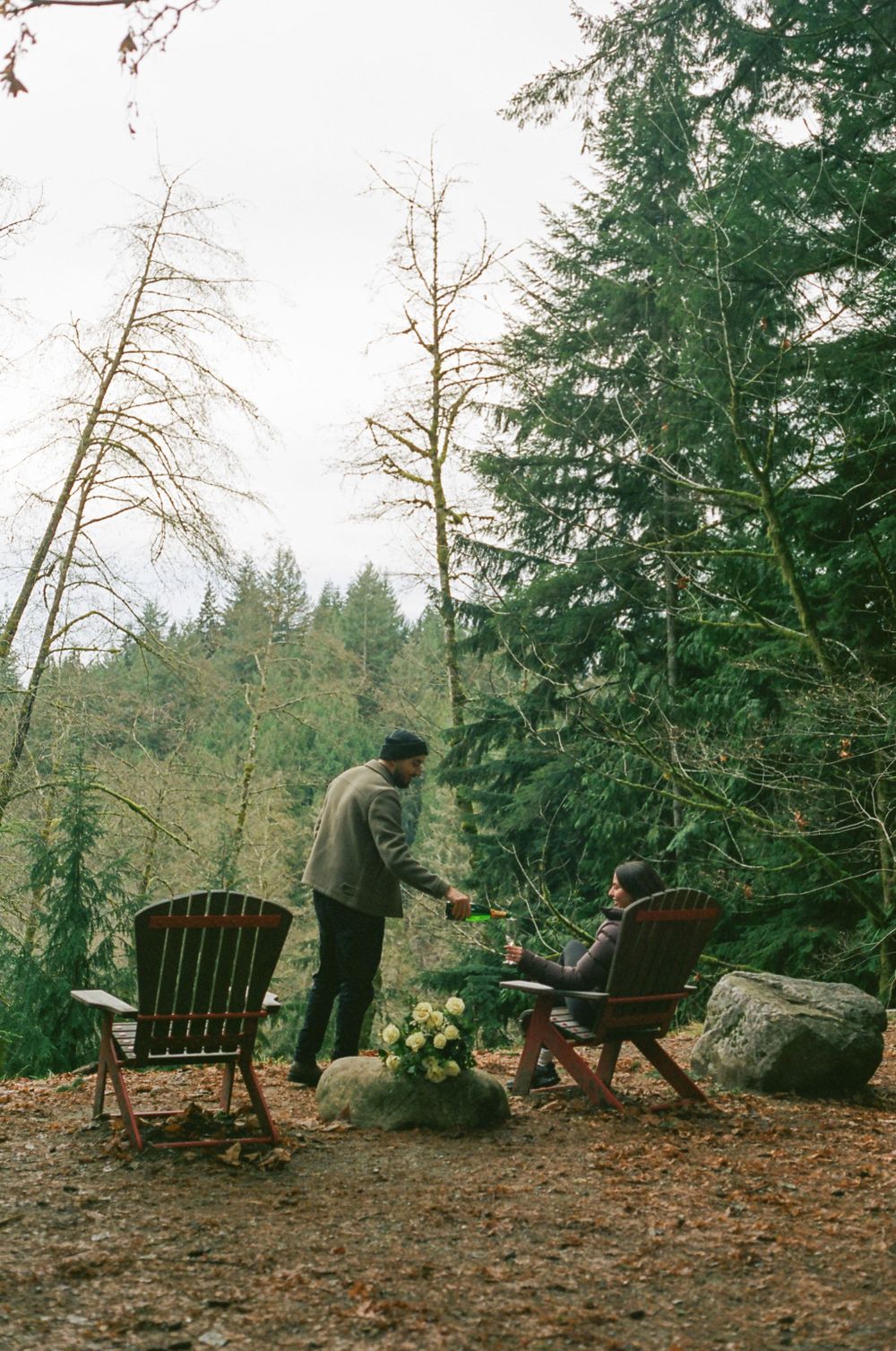 Wooden Adirondack chairs arranged in a peaceful forest setting with fallen leaves.