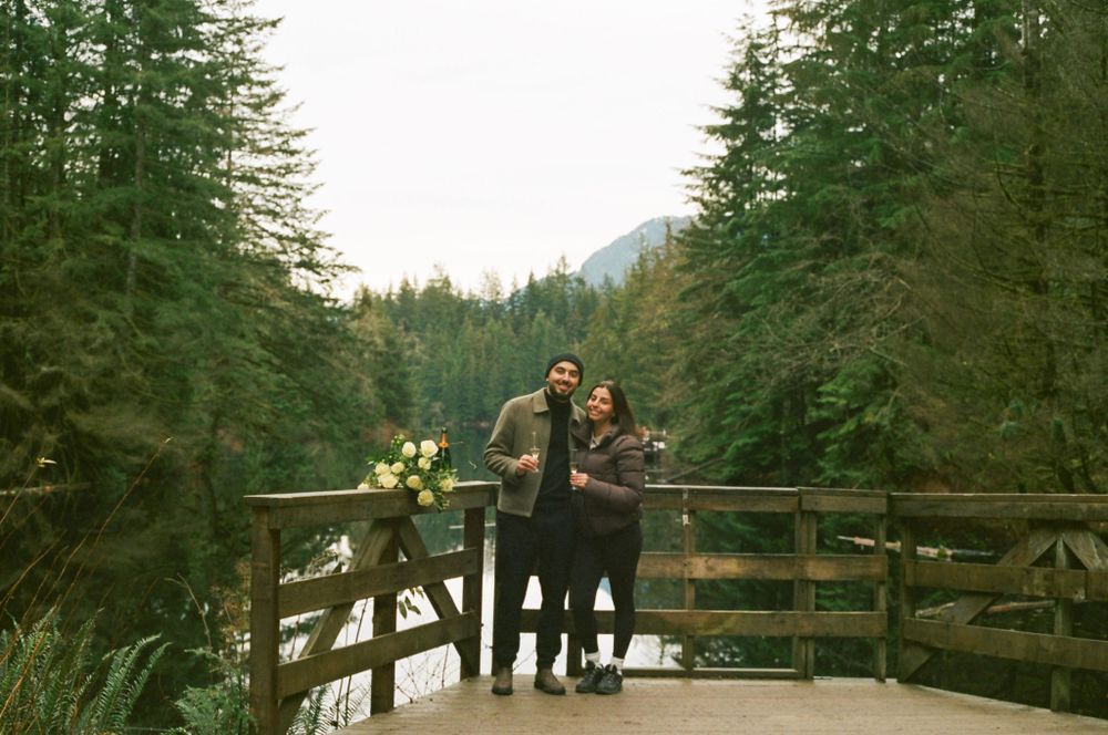 A long-distance view of two people celebrating on a wooden observation deck surrounded by evergreen forest.