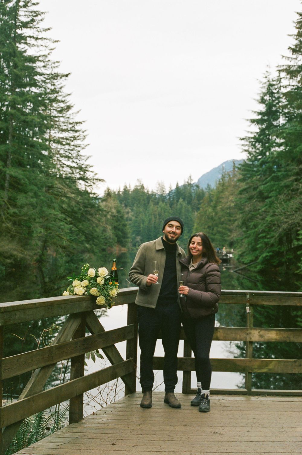 A romantic outdoor moment with two people standing on a wooden deck overlooking a misty forest landscape.