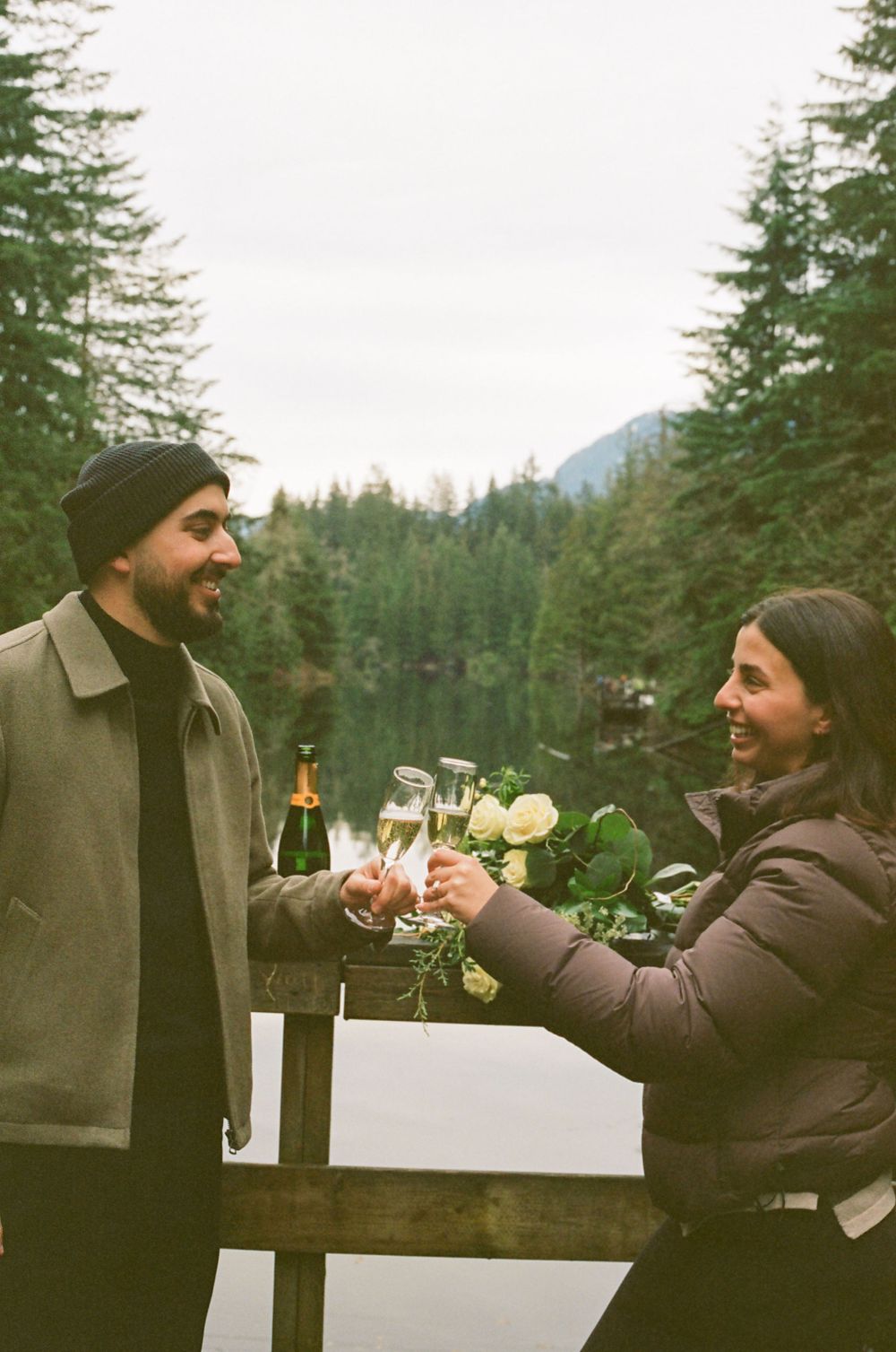 A couple sharing a celebratory toast on a foggy mountain deck with pine trees in the background.