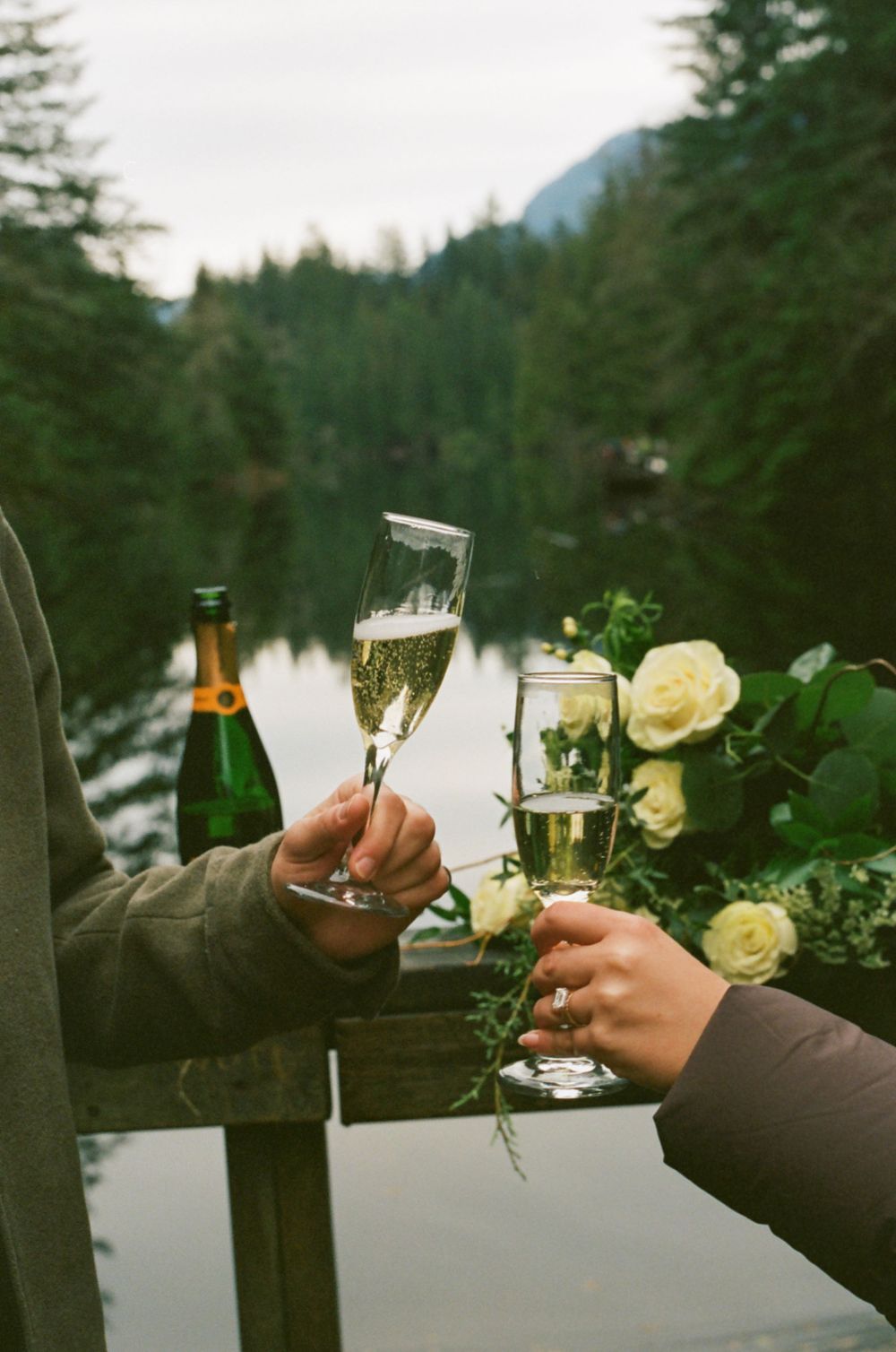 Champagne toast with flowers decorating wooden deck railing by mountain lake.