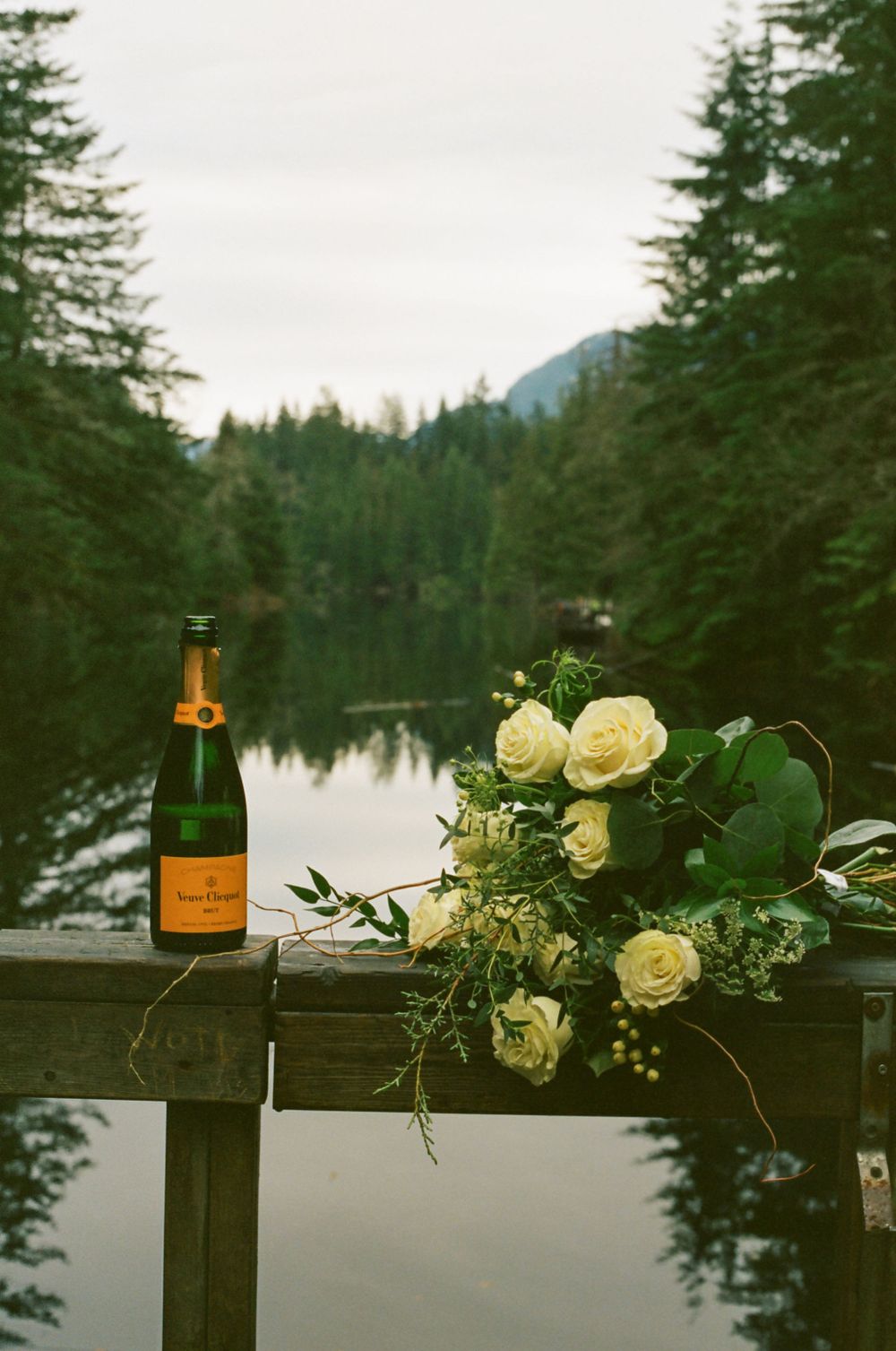 White roses and champagne bottles arranged on wooden railing with lake view.