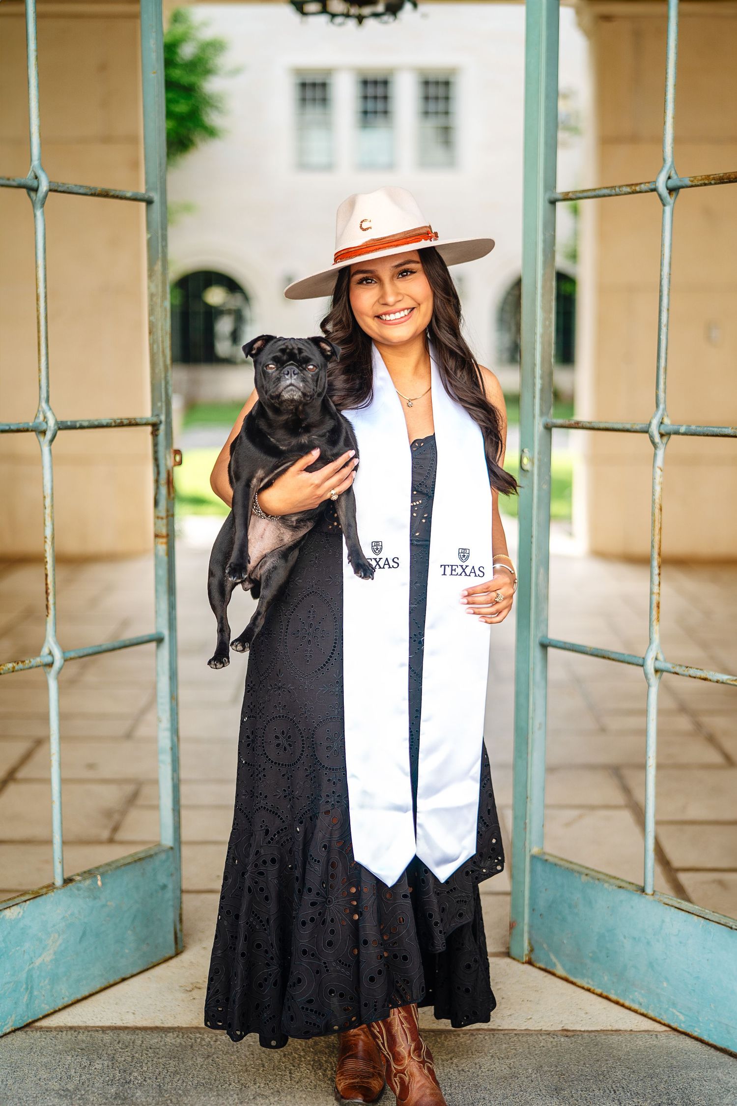Graduate in black dress and hat holding small black dog in architectural doorway.