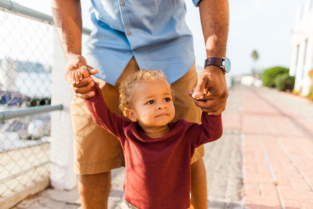 toddler holding both of dad's hands as he walks along a street.