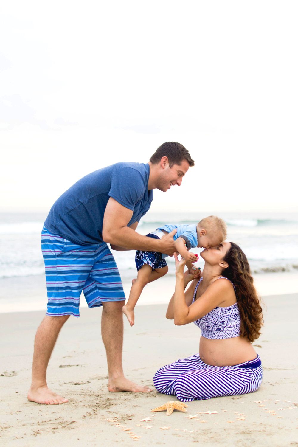 family at the beach. dad leaning their toddler down so the toddler can give pregnant mom a kiss