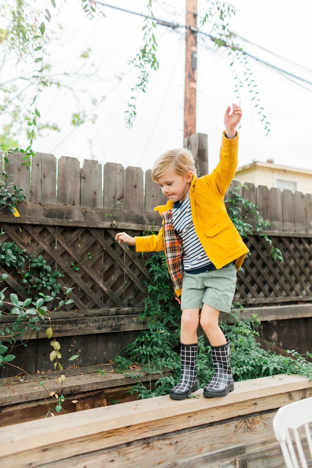 little boy in yellow rain coat balancing on a ledge in front of a fence