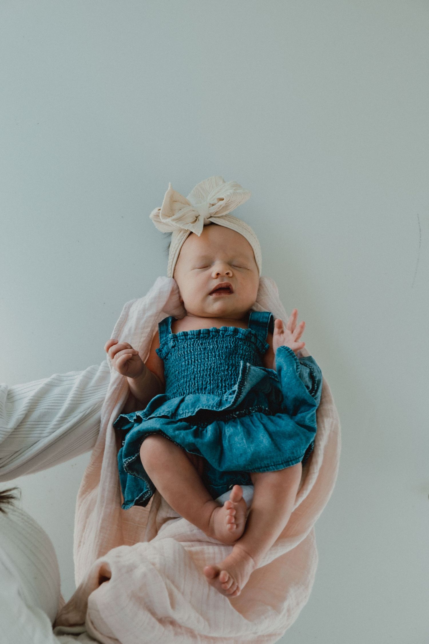 Sleeping baby wearing teal blue dress and white bow headband resting on soft pink blanket against light background.