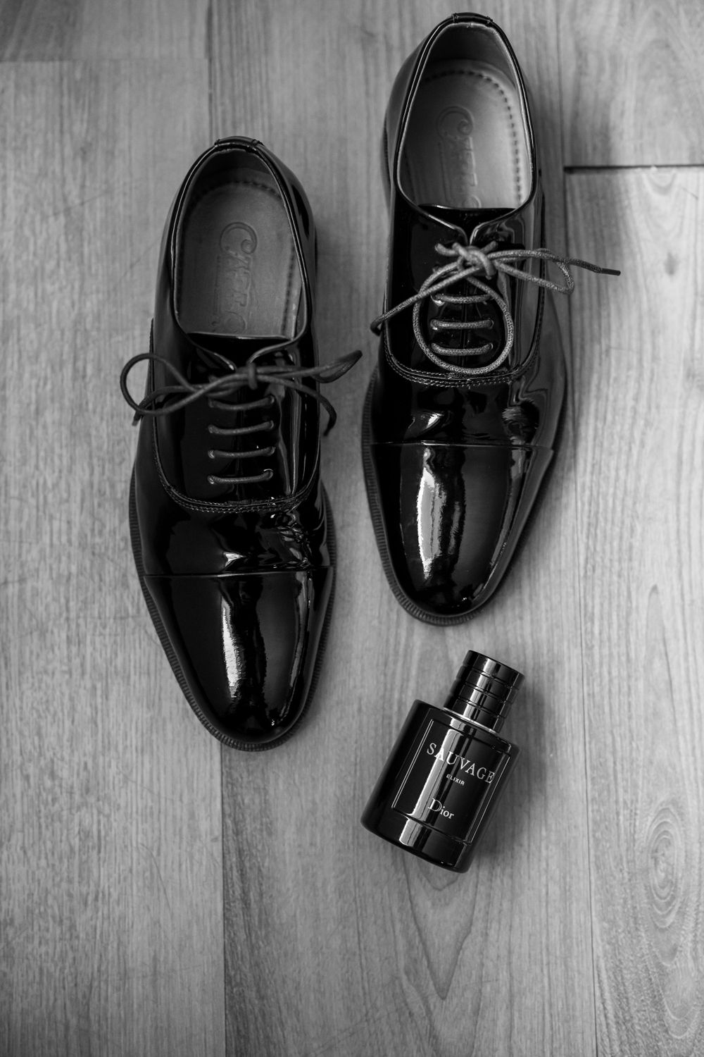 Elegant black patent leather dress shoes with a bottle of cologne on wooden floor.
