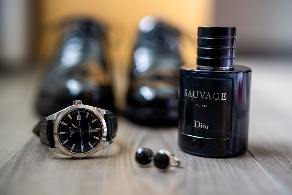 Luxury accessories including dress shoes, Dior Sauvage perfume, watch and cufflinks on wooden surface.