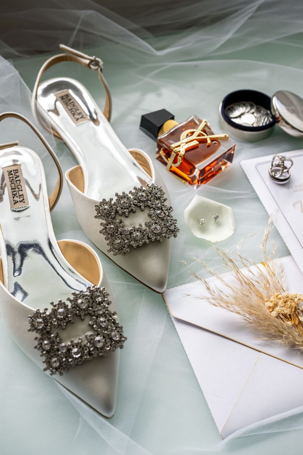 Elegant bridal shoes with crystal embellishments displayed with perfume and wedding accessories on tulle fabric.