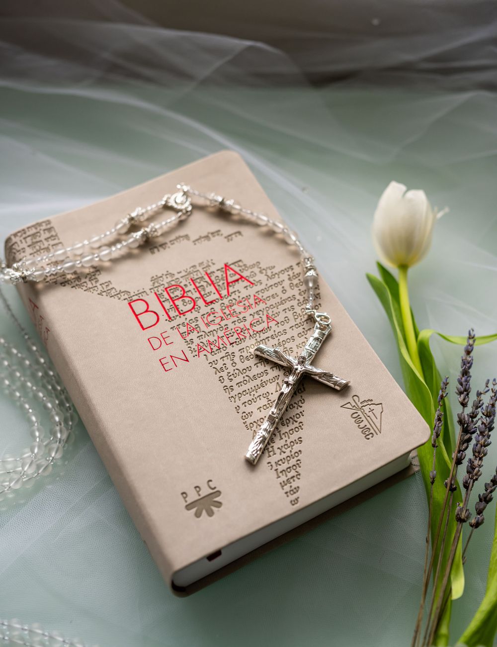 Bible with rosary beads and white tulip on light fabric background.