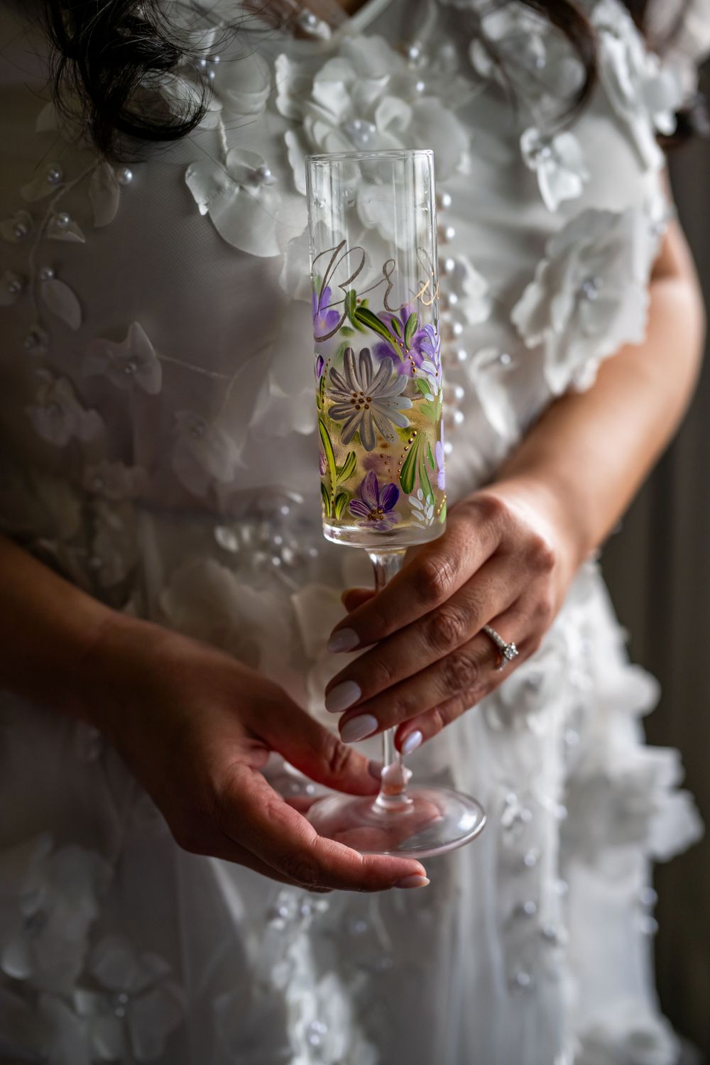 Hands holding decorative champagne flute with painted purple flowers against white dress.