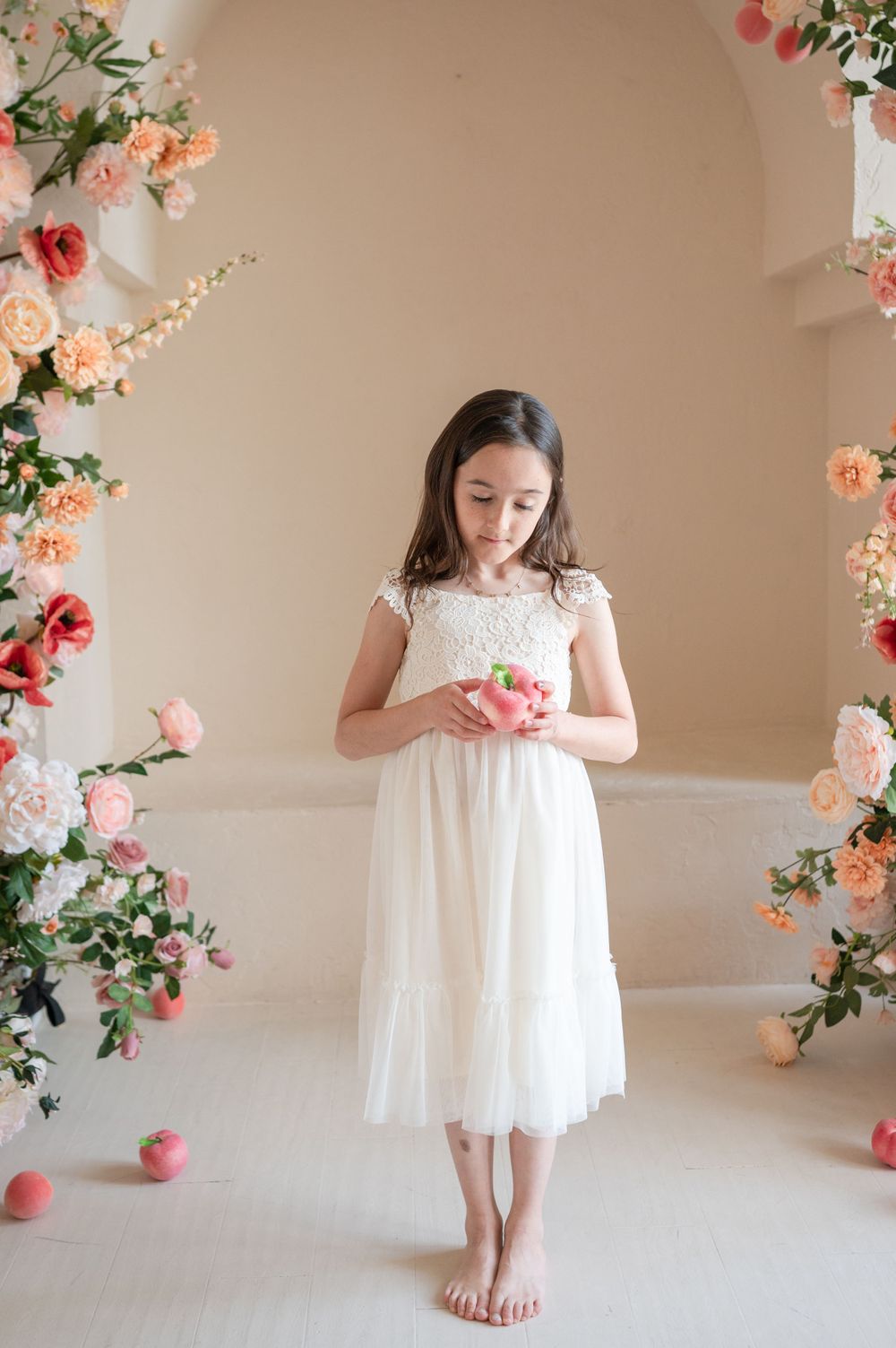 A young person in white stands contemplatively under a rose-covered arch holding a pink flower.