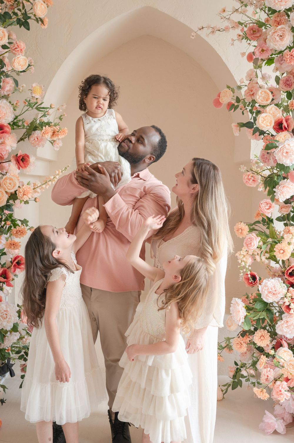 Family portrait under floral arch with everyone wearing white and pink coordinated outfits.